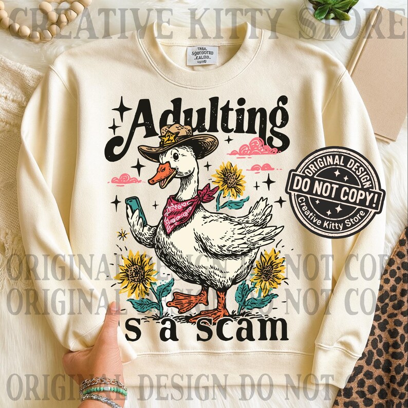 May include: Cream sweatshirt featuring a cartoon goose in a cowboy hat and bandana, holding a phone. The text "Adulting's a scam" is displayed with sunflowers and clouds. A Creative Kitty Store design.