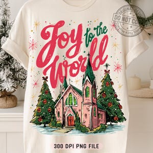 May include: Cream-colored t-shirt with a Christmas-themed graphic. The design features the text "Joy to the World" in red and green, a pink church, and two decorated Christmas trees. The bottom of the shirt has the text "300 DPI PNG FILE".