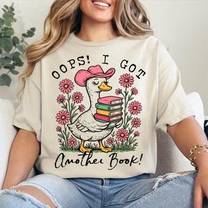 May include: A white t-shirt with a pink duck wearing a pink cowboy hat and holding a stack of books. The text on the shirt reads "Oops! I got another book!"