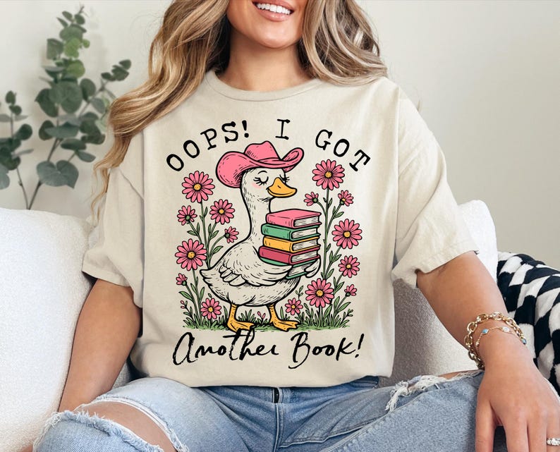 May include: A white t-shirt with a pink duck wearing a pink cowboy hat and holding a stack of books. The text on the shirt reads "Oops! I got another book!"