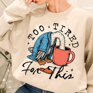 Op de afbeelding: Een cr&egrave;mekleurige trui met een afbeelding van een eend die een blauwe badjas draagt en koffie drinkt uit een rode mok. De tekst "Too Tired For This" staat op de trui gedrukt.