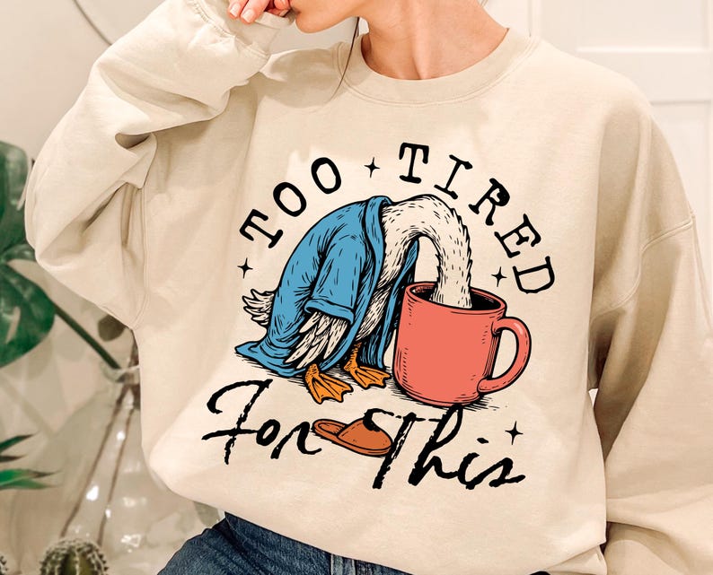 Op de afbeelding: Een cr&egrave;mekleurige trui met een afbeelding van een eend die een blauwe badjas draagt en koffie drinkt uit een rode mok. De tekst "Too Tired For This" staat op de trui gedrukt.