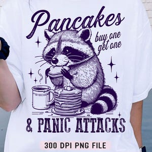 May include: A white t-shirt featuring a black and white illustration of a raccoon eating pancakes. The text "Pancakes buy one get one & Panic Attacks" is printed on the shirt.  300 DPI PNG FILE.
