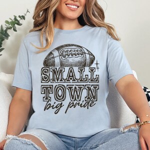 May include: A light blue t-shirt with a vintage-style football graphic and the text "SMALL TOWN big pride".