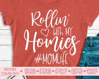 May include: A red t-shirt with the words "Rollin' with my Homies" in white script, a heart graphic, and the hashtag #MOM LIFE. The shirt is paired with blue jeans.
