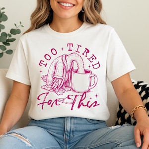 Op de afbeelding: Wit T-shirt met een roze afbeelding van een cartoon eend die een badjas draagt en uit een mok drinkt. De tekst "Too Tired For This" staat op het shirt gedrukt.