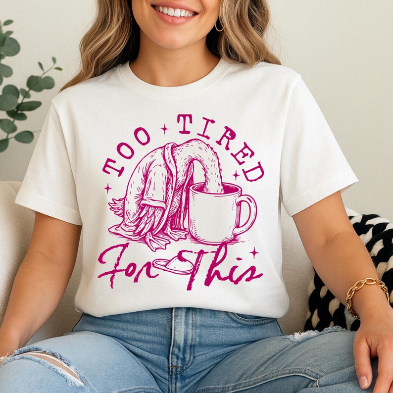 Op de afbeelding: Wit T-shirt met een roze afbeelding van een cartoon eend die een badjas draagt en uit een mok drinkt. De tekst "Too Tired For This" staat op het shirt gedrukt.