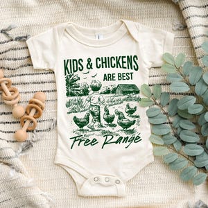 K&ouml;nnte beinhalten: Ein wei&szlig;er Baby-Strampler mit einer gr&uuml;nen Grafik eines Kindes und H&uuml;hnern. Der Text lautet "Kids & Chickens Are Best Free Range".