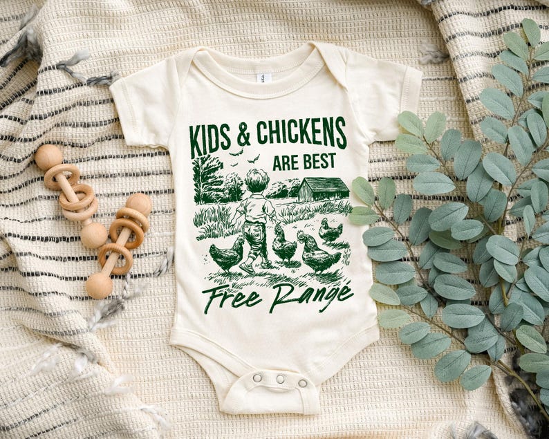 K&ouml;nnte beinhalten: Ein wei&szlig;er Baby-Strampler mit einer gr&uuml;nen Grafik eines Kindes und H&uuml;hnern. Der Text lautet "Kids & Chickens Are Best Free Range".