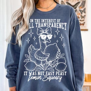 May include: A dark blue sweatshirt with a white graphic of a duck wearing sunglasses and holding lemons. The text on the sweatshirt reads "In the interest of full transparency it was not easy peasy lemon squeezy."