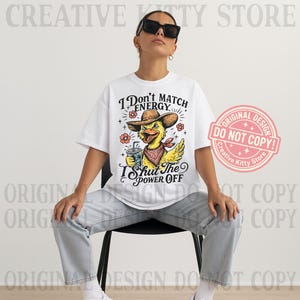 May include: White t-shirt with a graphic of a yellow duck wearing a cowboy hat and bandana, holding a drink. The text on the shirt reads "I Don't Match Energy, I Shut The Power Off." A pink and white "Original Design" sticker is on the right.