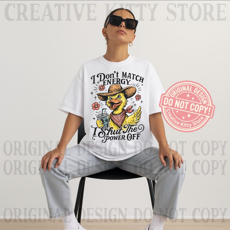 May include: White t-shirt with a graphic of a yellow duck wearing a cowboy hat and bandana, holding a drink. The text on the shirt reads "I Don't Match Energy, I Shut The Power Off." A pink and white "Original Design" sticker is on the right.