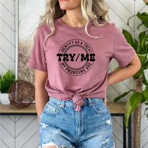 Puede incluir: Una camiseta color malva con un gr&aacute;fico circular negro que dice "Identify as a treat, Try/Me, My pronouns are".