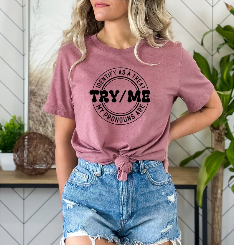 Puede incluir: Una camiseta color malva con un gr&aacute;fico circular negro que dice "Identify as a treat, Try/Me, My pronouns are".
