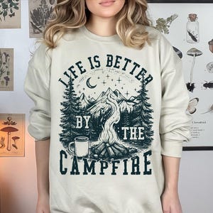 Puede incluir: Sudadera de color crema con un dise&ntilde;o gr&aacute;fico que presenta una escena de fogata. El dise&ntilde;o incluye una cordillera, &aacute;rboles, una luna creciente y el texto "LIFE IS BETTER BY THE CAMPFIRE" en verde oscuro.