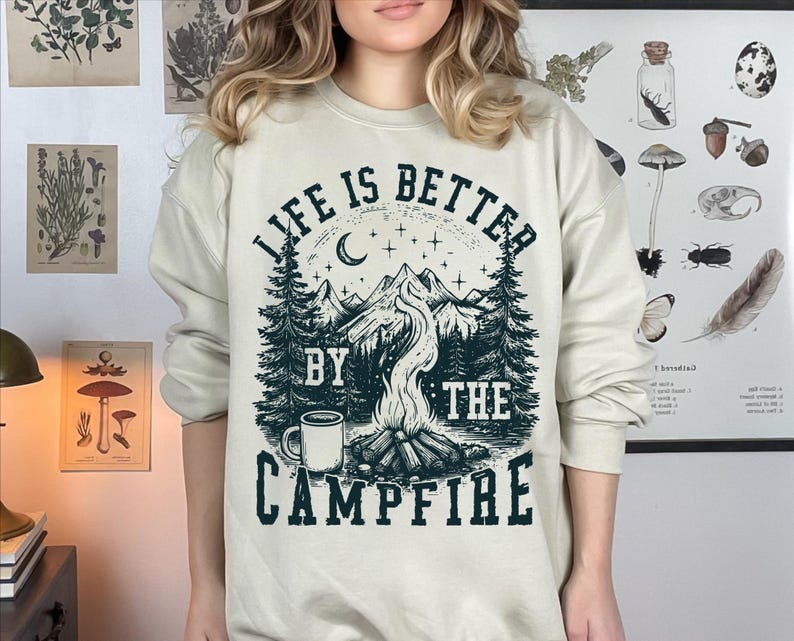 Puede incluir: Sudadera de color crema con un dise&ntilde;o gr&aacute;fico que presenta una escena de fogata. El dise&ntilde;o incluye una cordillera, &aacute;rboles, una luna creciente y el texto "LIFE IS BETTER BY THE CAMPFIRE" en verde oscuro.