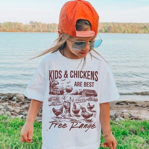 K&ouml;nnte beinhalten: Wei&szlig;es T-Shirt mit einer braunen Grafik von Kindern und H&uuml;hnern. Der Text auf dem Shirt lautet "Kids & Chickens Are Best Free Range".