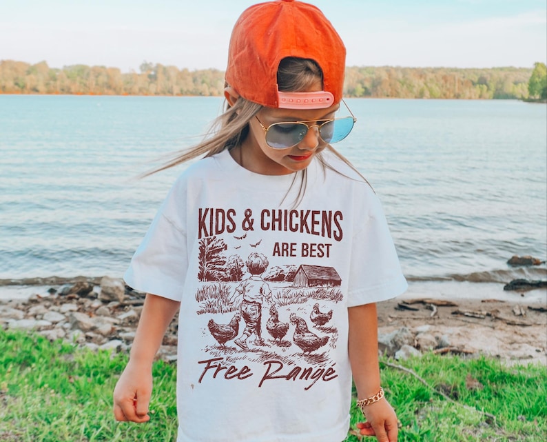 K&ouml;nnte beinhalten: Wei&szlig;es T-Shirt mit einer braunen Grafik von Kindern und H&uuml;hnern. Der Text auf dem Shirt lautet "Kids & Chickens Are Best Free Range".