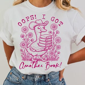 May include: White t-shirt with a pink graphic of a duck wearing a cowboy hat and carrying a stack of books. The text "Oops! I got another book!" is printed on the shirt.