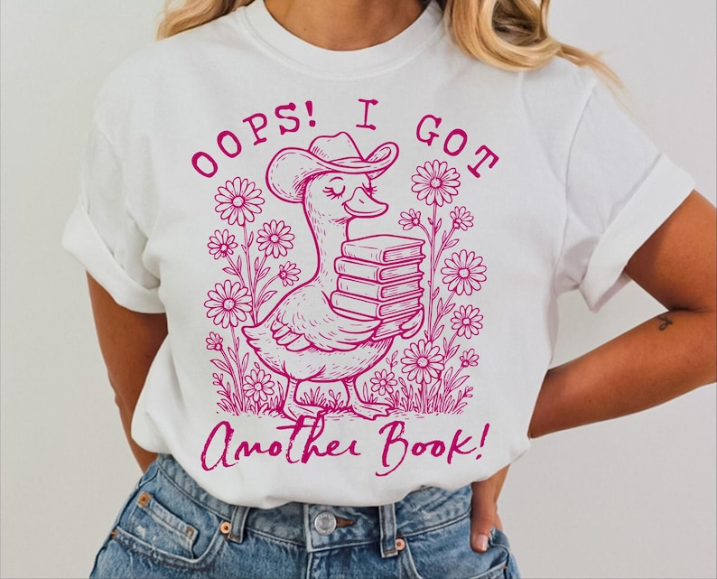 May include: White t-shirt with a pink graphic of a duck wearing a cowboy hat and carrying a stack of books. The text "Oops! I got another book!" is printed on the shirt.