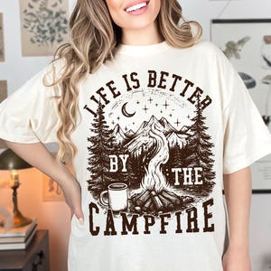 Puede incluir: Camiseta color crema con un gr&aacute;fico marr&oacute;n que dice "LIFE IS BETTER BY THE CAMPFIRE". El dise&ntilde;o presenta una fogata, monta&ntilde;as, &aacute;rboles, una luna creciente, estrellas y una taza de caf&eacute;.