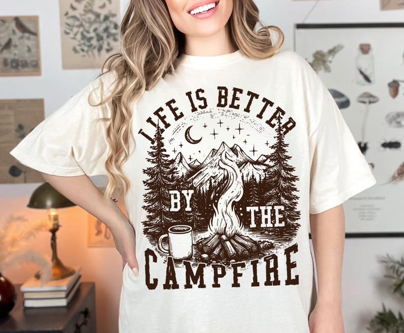 Puede incluir: Camiseta color crema con un gr&aacute;fico marr&oacute;n que dice "LIFE IS BETTER BY THE CAMPFIRE". El dise&ntilde;o presenta una fogata, monta&ntilde;as, &aacute;rboles, una luna creciente, estrellas y una taza de caf&eacute;.