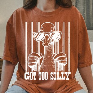 Pu&ograve; includere: Una maglietta color ruggine con una grafica bianca di un'anatra con occhiali da sole e il testo "Got Too Silly".