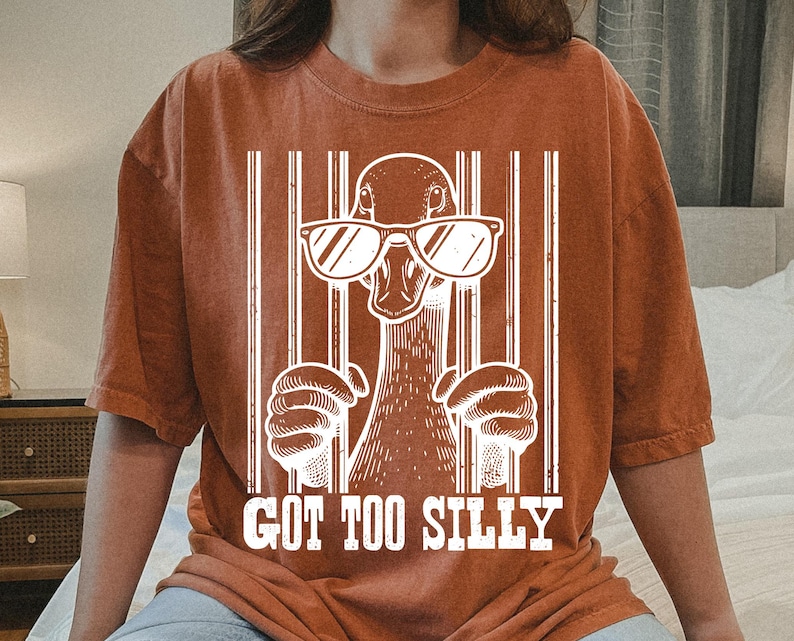 Pu&ograve; includere: Una maglietta color ruggine con una grafica bianca di un'anatra con occhiali da sole e il testo "Got Too Silly".