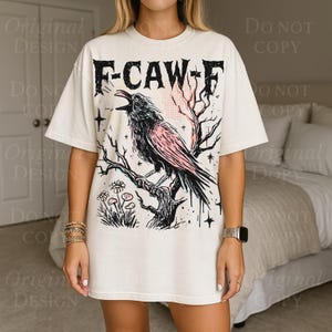 May include: Cream-colored t-shirt featuring a black and pink crow perched on a branch, with the text "F-CAW-F" above. The design includes a tree, flowers, and stars. The shirt has short sleeves and a relaxed fit.