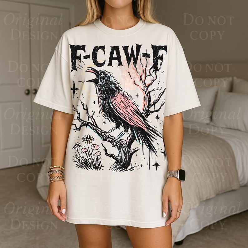 May include: Cream-colored t-shirt featuring a black and pink crow perched on a branch, with the text "F-CAW-F" above. The design includes a tree, flowers, and stars. The shirt has short sleeves and a relaxed fit.