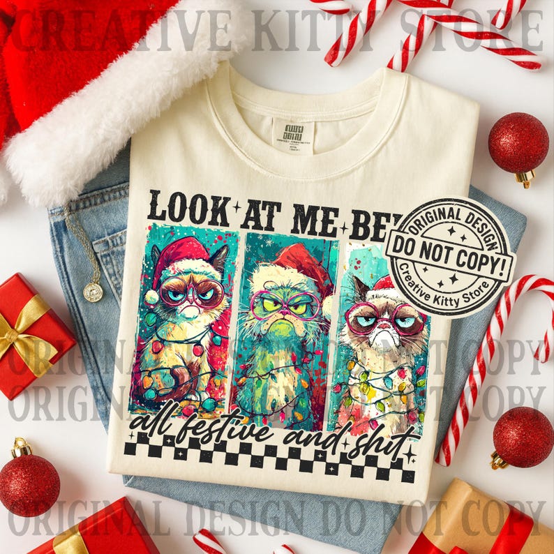 May include: Cream-colored t-shirt featuring a graphic of three grumpy cats wearing Santa hats and Christmas lights, with the text "LOOK AT ME-BE" and "all festive and shit". The shirt is surrounded by holiday decorations.
