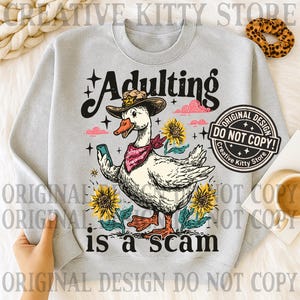 May include: A pale grey sweatshirt featuring a cartoon goose in a cowboy hat and bandana, holding a phone. The text reads "Adulting is a scam." The design includes sunflowers and stars.