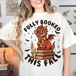 K&ouml;nnte beinhalten: Cremefarbenes T-Shirt mit einem Cartoon-Drachen, der eine Brille und einen Schal tr&auml;gt und auf B&uuml;chern sitzt. Der Text "FULLY BOOKED THIS FALL" befindet sich &uuml;ber und unter dem Drachen. Flederm&auml;use, Sterne und Kerzen vervollst&auml;ndigen das Design.