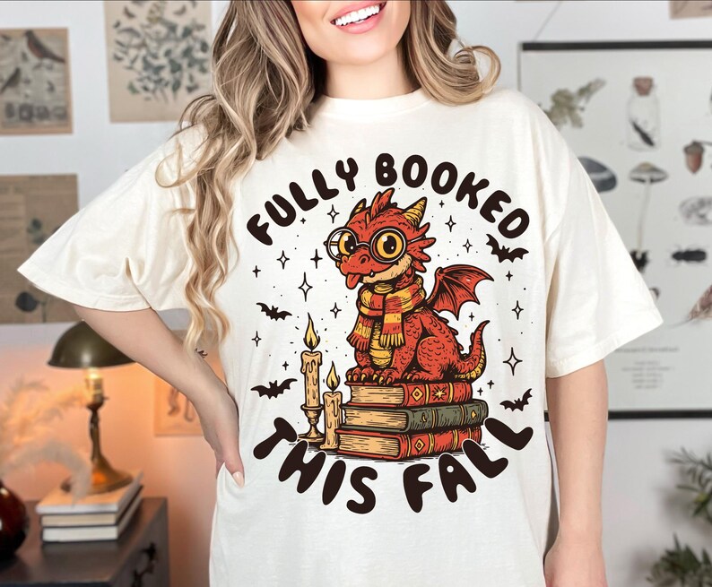 K&ouml;nnte beinhalten: Cremefarbenes T-Shirt mit einem Cartoon-Drachen, der eine Brille und einen Schal tr&auml;gt und auf B&uuml;chern sitzt. Der Text "FULLY BOOKED THIS FALL" befindet sich &uuml;ber und unter dem Drachen. Flederm&auml;use, Sterne und Kerzen vervollst&auml;ndigen das Design.