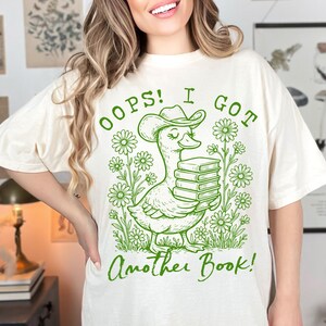May include: A white t-shirt with a green graphic of a duck wearing a cowboy hat and holding a stack of books. The text "Oops! I got another book!" is printed on the shirt.