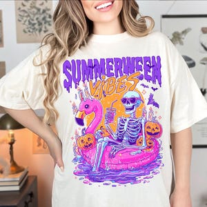 May include: A cream-coloured t-shirt with a vibrant graphic. The design shows a skeleton in sunglasses, lounging on a pink flamingo float, with the words "Summerween Vibes" in purple and orange. Pumpkins and bats are also featured in the design.