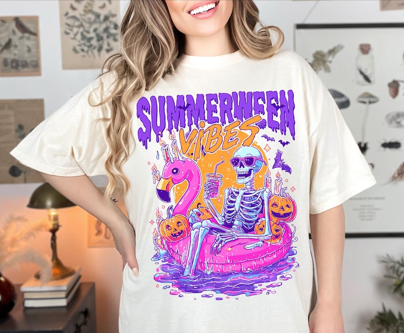 May include: A cream-coloured t-shirt with a vibrant graphic. The design shows a skeleton in sunglasses, lounging on a pink flamingo float, with the words "Summerween Vibes" in purple and orange. Pumpkins and bats are also featured in the design.