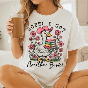 May include: White t-shirt with a cartoon duck wearing a cowboy hat and holding a stack of books. The text on the shirt reads "Oops! I got another book!"