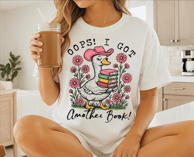 May include: White t-shirt with a cartoon duck wearing a cowboy hat and holding a stack of books. The text on the shirt reads "Oops! I got another book!"