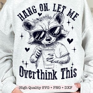 Puede incluir: Una sudadera gris con una ilustración en blanco y negro de un mapache con gafas de sol y sosteniendo un batido. El texto "HANG ON. LET ME OVERTHINK THIS" está impreso en la sudadera.
