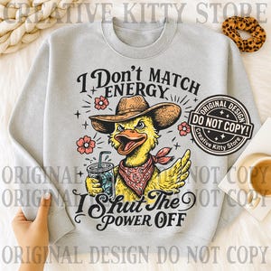 May include: A light gray sweatshirt featuring a cartoon duck wearing a cowboy hat and a red bandana, holding a drink. The text reads "I Don't Match Energy, I Shut The Power Off." The design includes flowers and stars.