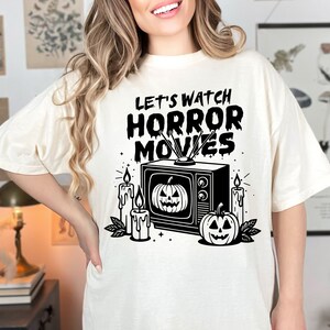 May include: A white t-shirt with a black graphic of a vintage television with the text "Let's Watch Horror Movies" and pumpkins and candles around it.