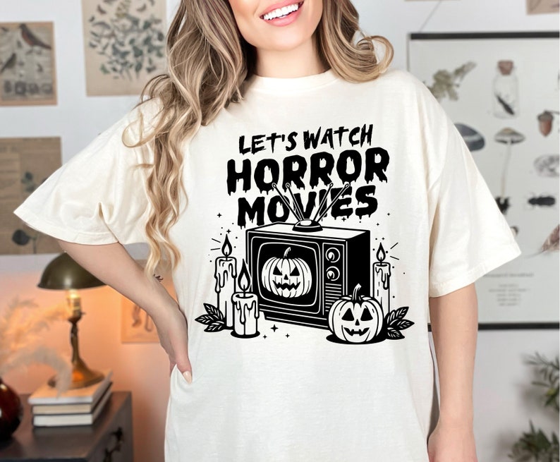 May include: A white t-shirt with a black graphic of a vintage television with the text "Let's Watch Horror Movies" and pumpkins and candles around it.