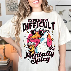 Pu&ograve; includere: T-shirt color crema con un'anatra dei cartoni animati con occhiali da sole e una bevanda. Il testo sulla maglietta recita "Expensive, Difficult & Mentally Spicy". Il design include accenti rosa, arancioni, neri e blu.