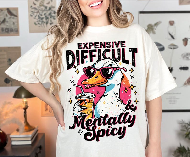 Pu&ograve; includere: T-shirt color crema con un'anatra dei cartoni animati con occhiali da sole e una bevanda. Il testo sulla maglietta recita "Expensive, Difficult & Mentally Spicy". Il design include accenti rosa, arancioni, neri e blu.