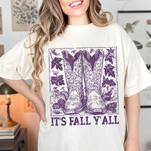 May include: Off-white t-shirt with a purple illustration of cowboy boots, autumn leaves, and pumpkins. The words "IT'S FALL Y'ALL" are printed beneath the image. The shirt has a loose fit.