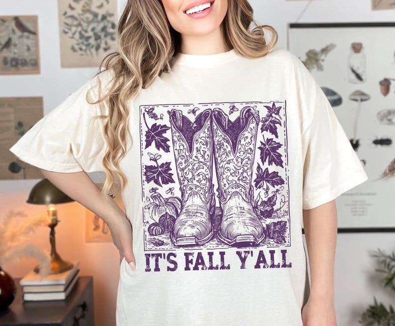 May include: Off-white t-shirt with a purple illustration of cowboy boots, autumn leaves, and pumpkins. The words "IT'S FALL Y'ALL" are printed beneath the image. The shirt has a loose fit.