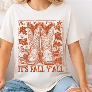 May include: Cream-coloured t-shirt with a rust-coloured graphic of cowboy boots, pumpkins, and autumn leaves. The text "IT'S FALL Y'ALL" is printed below. The shirt is made of a soft material.