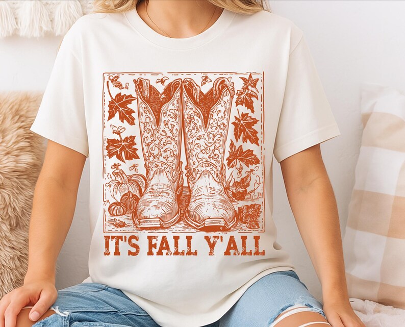 May include: Cream-coloured t-shirt with a rust-coloured graphic of cowboy boots, pumpkins, and autumn leaves. The text "IT'S FALL Y'ALL" is printed below. The shirt is made of a soft material.
