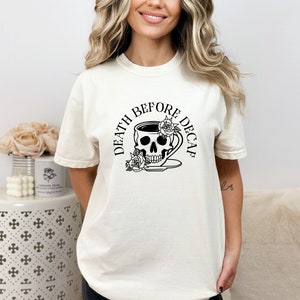 May include: A cream-coloured t-shirt with a black graphic. The graphic shows a skull-shaped coffee cup with roses and the text "DEATH BEFORE DECAF". The shirt has short sleeves and a relaxed fit.